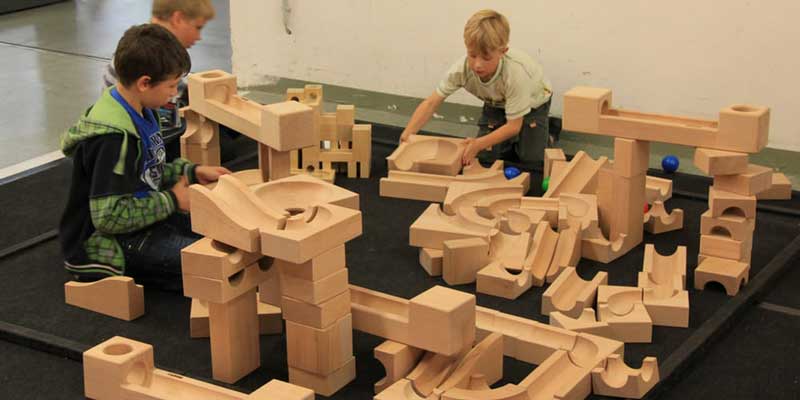 Zdjęcie: Trzej chłopcy bawią się, budując kulodrom z gigantycznych drewnianych klocków (producent - Kaden, seria XL). Na podłodze widać też olbrzymie drewniane kulki.